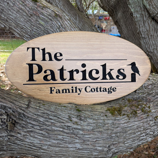 Personalized engraved wood oval sign 'The Patricks Family Cottage' with bird motif on wooden perch. Custom cabin address plaque or welcome sign with Established Date.