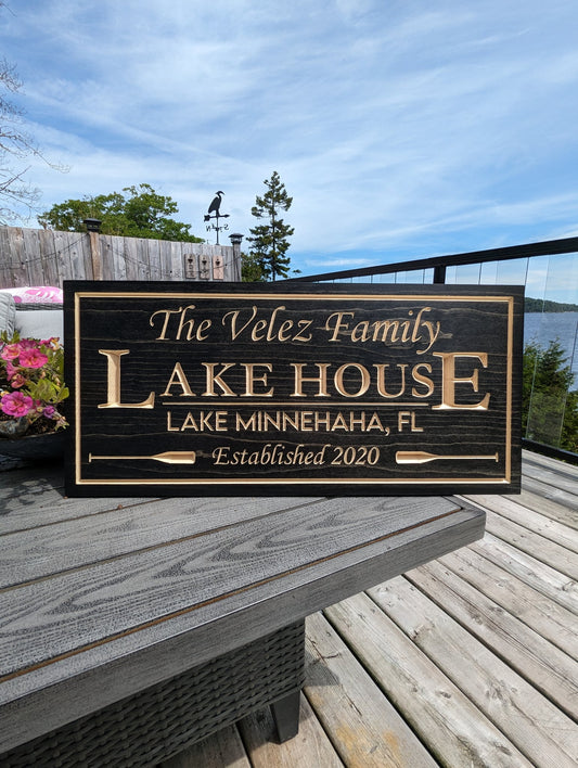 Personalized engraved lake house sign displayed on a wooden deck with a scenic lake view background. Custom cottage decor for outdoor living spaces.