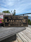 Personalized engraved lake house sign displayed on a wooden deck with a scenic lake view background. Custom cottage decor for outdoor living spaces.