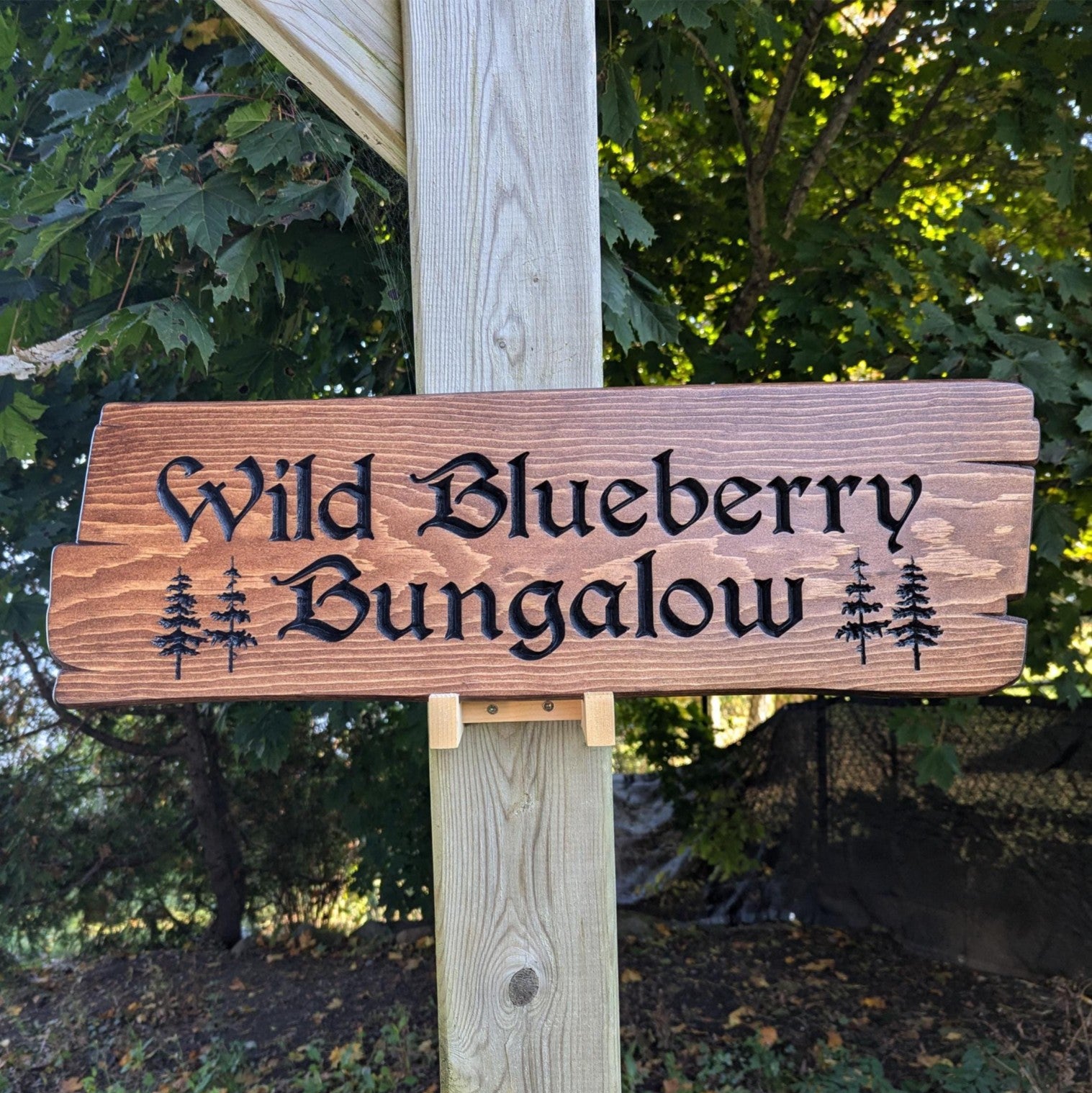 Custom engraved wood sign with 'Wild Blueberry Bungalow' text, mounted on a wooden post with rustic tree graphics and forest background. Personalized home decor.