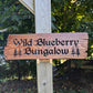 Custom engraved wood sign with 'Wild Blueberry Bungalow' text, mounted on a wooden post with rustic tree graphics and forest background. Personalized home decor.